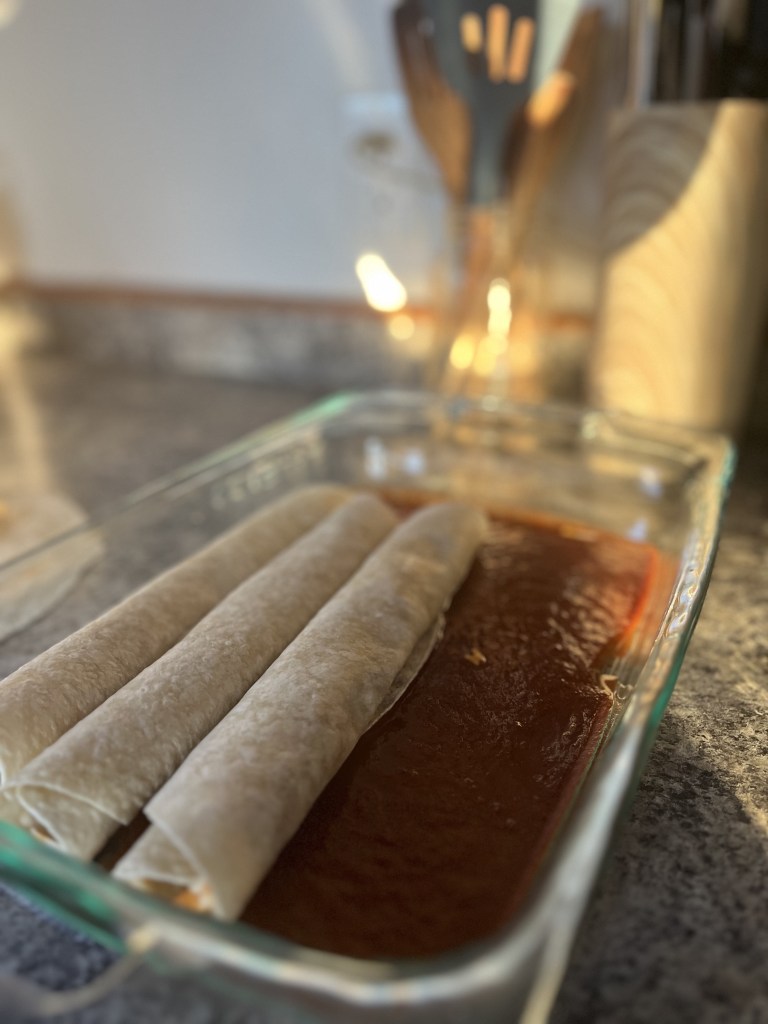 Three rolled tortillas on red sauce in a glass baking dish; kitchen utensils and warm lighting in the blurred background create a cozy mood.