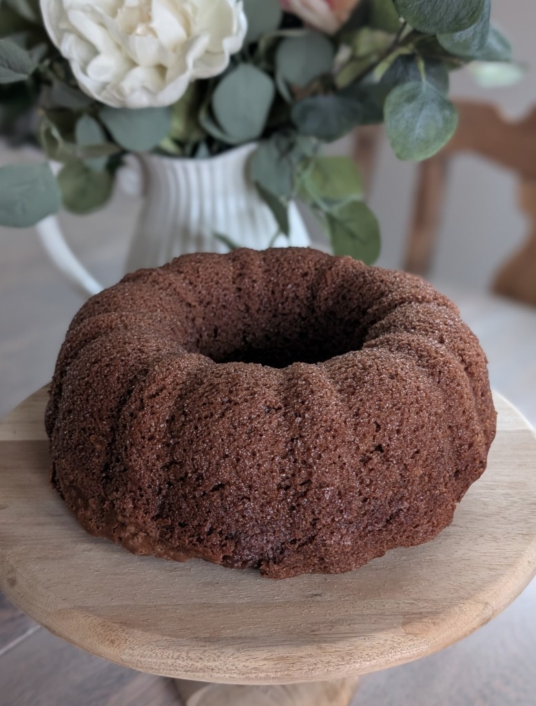 Chocolate bundt cake on a wooden stand with a blurred background showing a white vase of flowers. Cozy and inviting atmosphere.