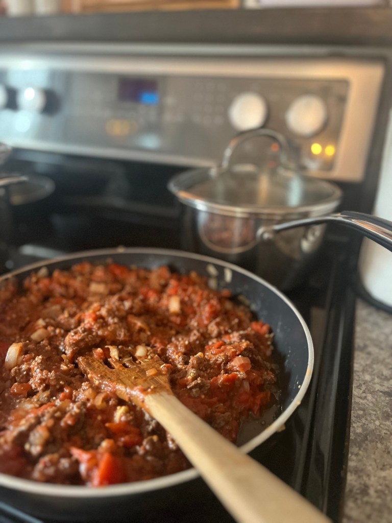 Skillet with ground meat and tomato sauce being stirred with a wooden spoon on a stove. Pot with lid in the background. Cozy kitchen scene.