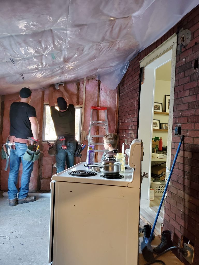 Two people work on wall insulation near a window in a room with exposed brick walls. A child watches by an old stove with pots.