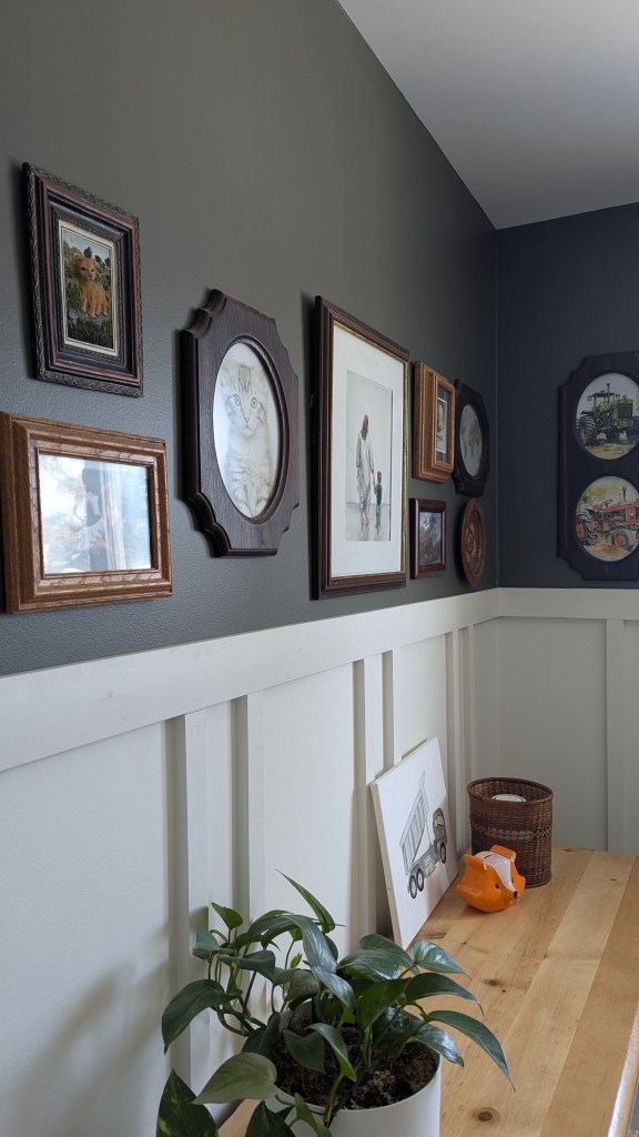 Gallery wall with framed art, a plant on a wooden table, a toy fox, and a drawing of a truck. Gray and white wall with wainscoting. Cozy setting.