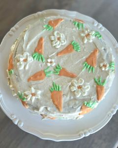 "Grandma's Carrot cake" on a white plate, topped with orange carrot designs and white flowers. The icing is creamy, creating a homemade feel.