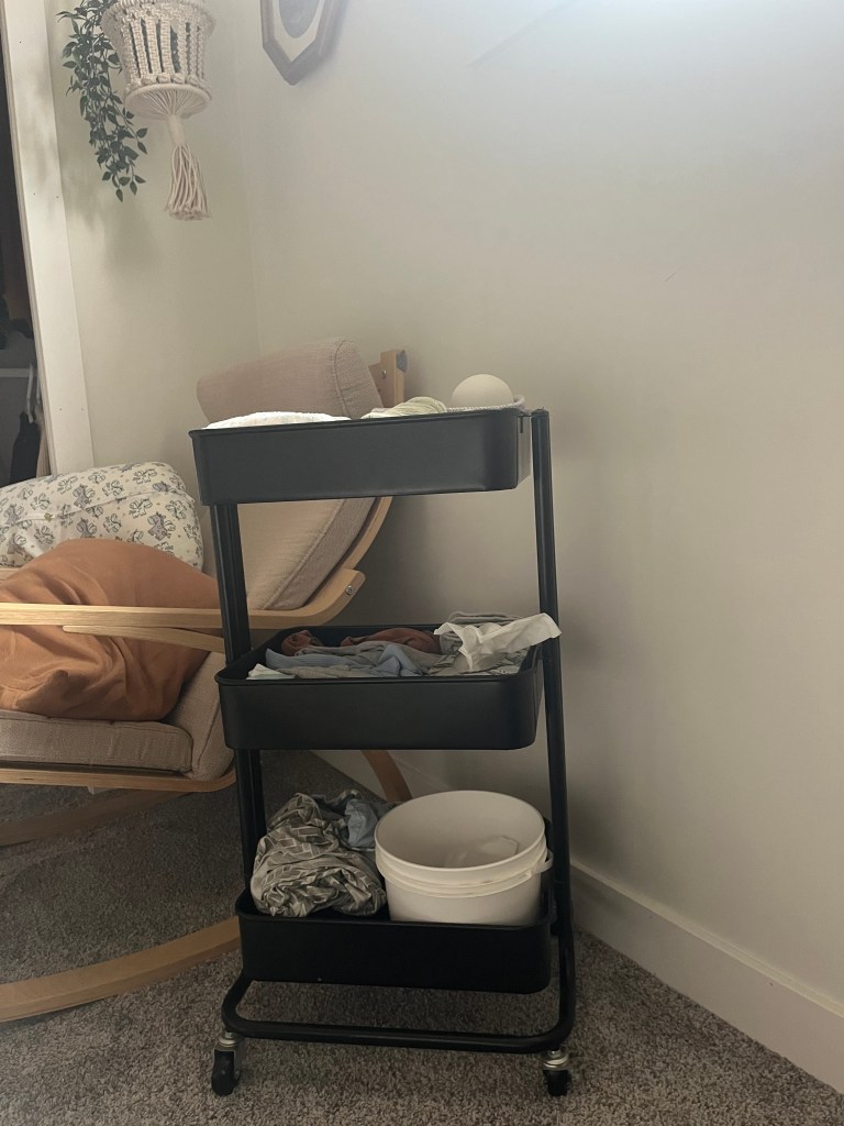 Black rolling cart with clothes and bucket next to a beige rocking chair with brown blanket. Macrame plant hanger on the wall. Cozy setting.