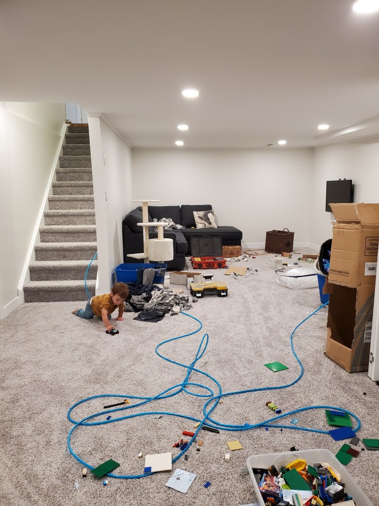 Child playing with toy car in messy basement with scattered Lego bricks and blue cables. Gray carpet, cardboard boxes, and black sofa.