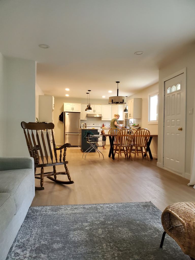 Open kitchen and dining area with wooden furniture. Person cooking at counter, cozy atmosphere, light wood floors, and modern lighting.