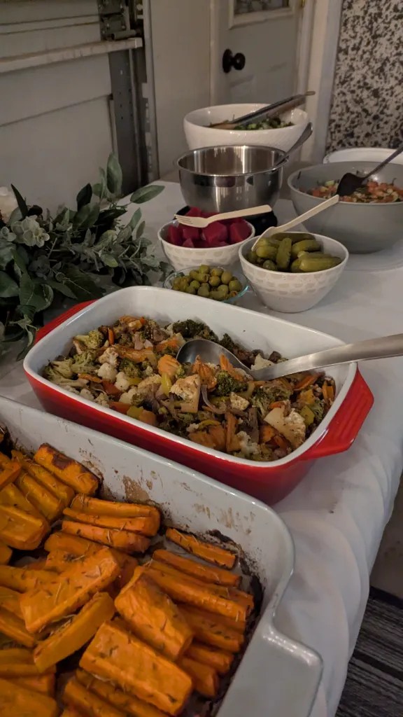 Buffet table with roasted carrots, mixed vegetables in red dish, pickles, and olives. White tablecloth, indoor setting, rustic decor.