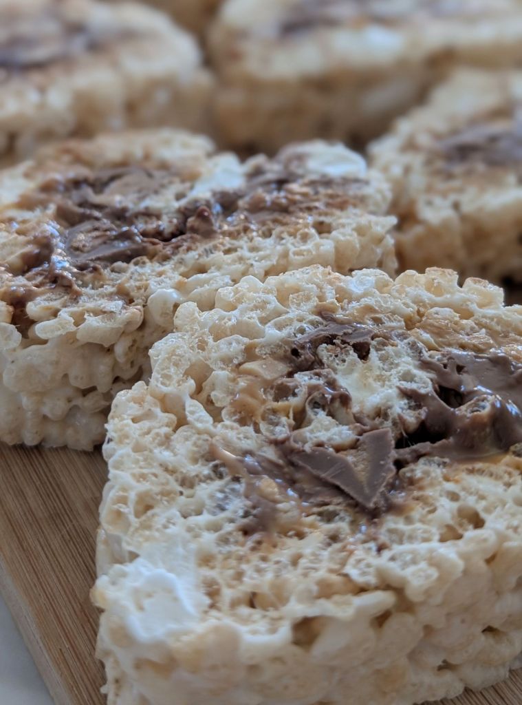 Close-up of Rice Krispies treats with chocolate swirls on a wooden board. The mood is indulgent. Background is softly blurred.