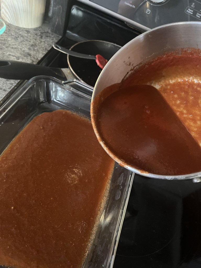 Pouring red sauce from a pot into a glass dish on a stovetop. Gray countertop and skillet in background, creating a cozy kitchen scene.