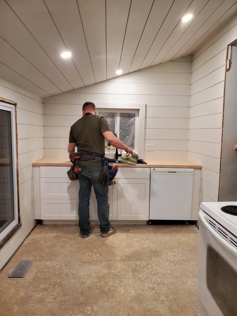 Worker in a toolbelt installs kitchen countertop in a white, modern kitchen with shiplap walls and recessed lighting. Bright, tidy space.