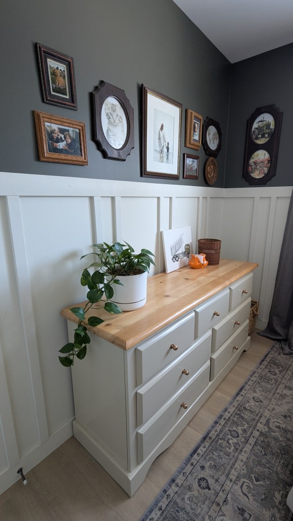 White dresser with a wooden top, potted plant, and framed art in a room with dark walls and wainscoting. Cozy, decorative setting.