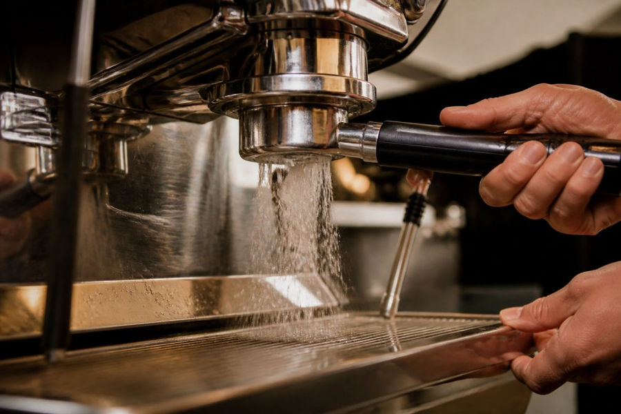 Professional barista's hands operating espresso machine group head valve during backflushing procedure, water spraying from portafilter basket, stainless steel machine visible in background with warm lighting