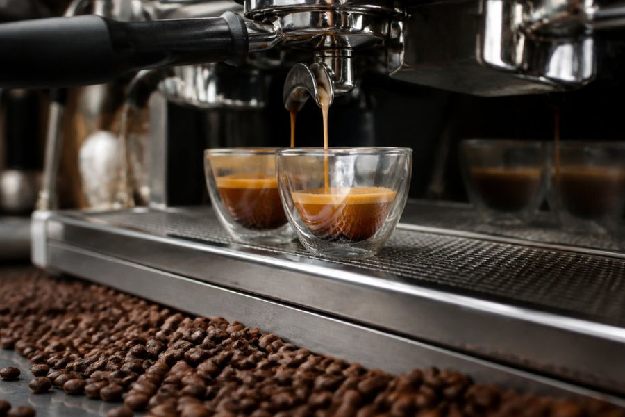 Close-up of espresso shots being pulled from a commercial espresso machine, showing rich golden crema formation and shot consistency, with whole espresso beans scattered in the foreground on a stainless steel counter