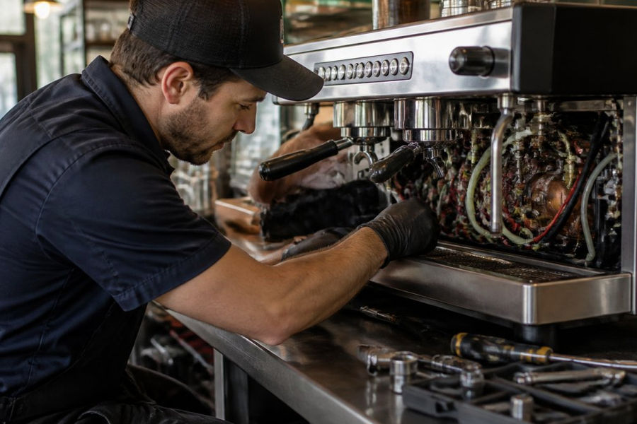 Professional technician in work gear performing diagnostic checks on a commercial espresso machine in a busy cafe setting, examining internal components with specialized tools