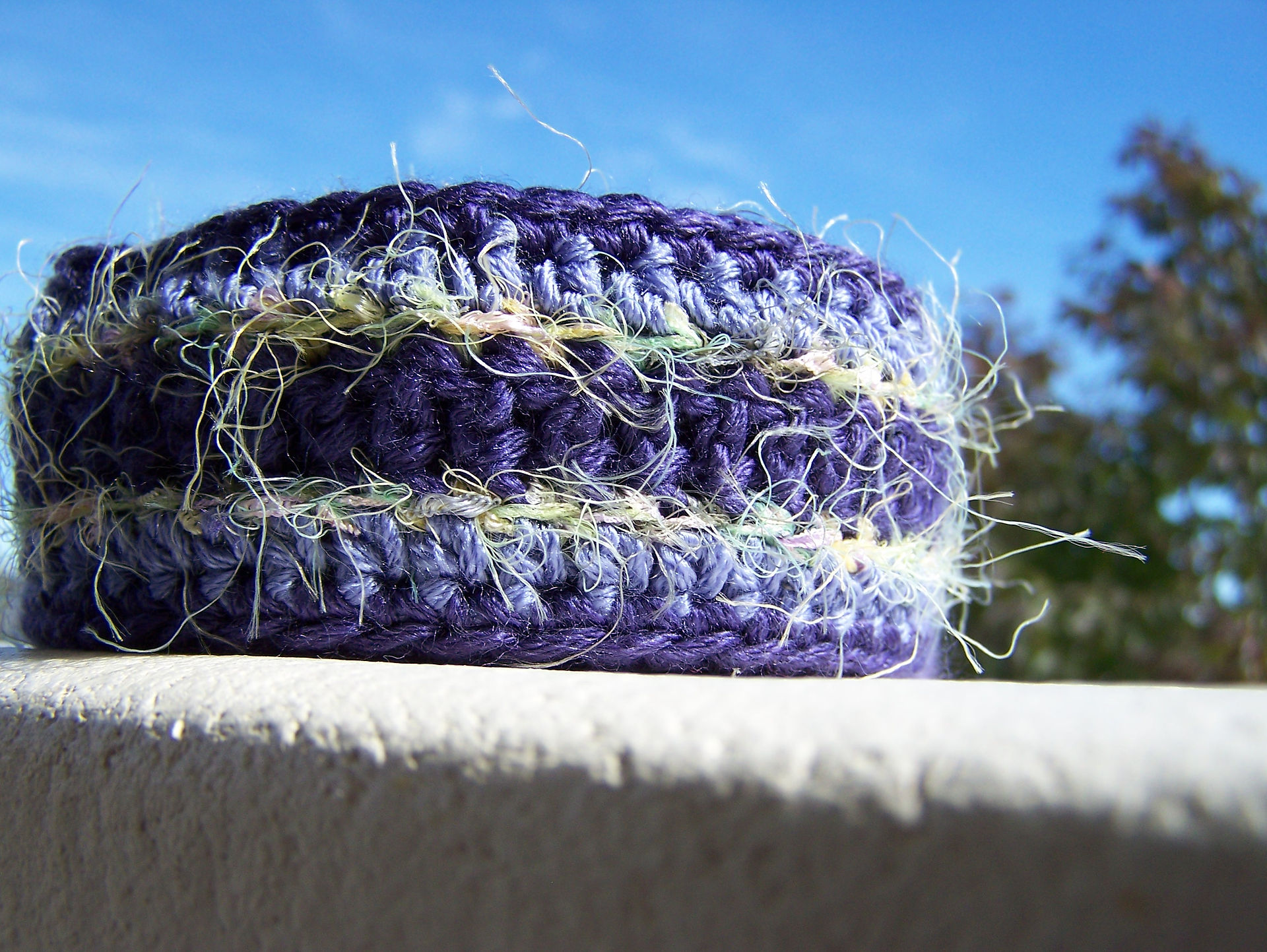 Purple striped handmade crocheted skinny scarf with fuzzy yellow and green stripes rolled up and sitting on a porch railing.
