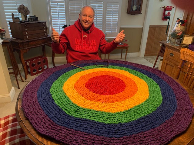 Rugby Jim sitting at a table with two thumbs up after just completing his largest rag rug to date at 49 inches in a rainbow of colors.
