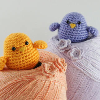 Yellow and lilac colored crochet bird rattles sitting inside color matching cones of thread with two crocheted flowers.