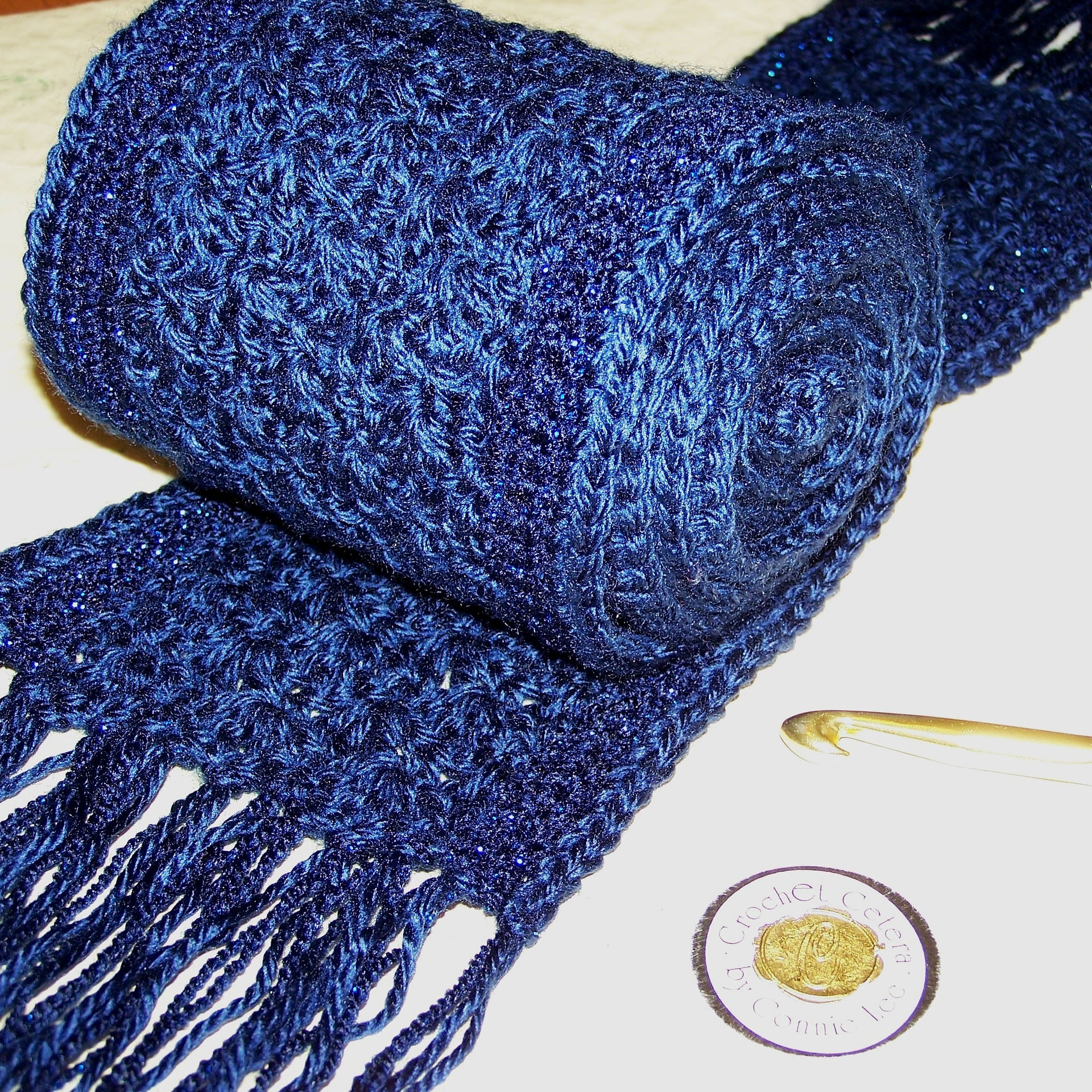 Dark blue crocheted scarf with fringe rolled up on a table next to a gold crochet hook.