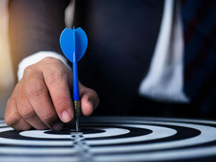 Man in a suit's hand placing a blue dart on a black-and-white dartboard, focusing on the bullseye. The setting conveys precision.