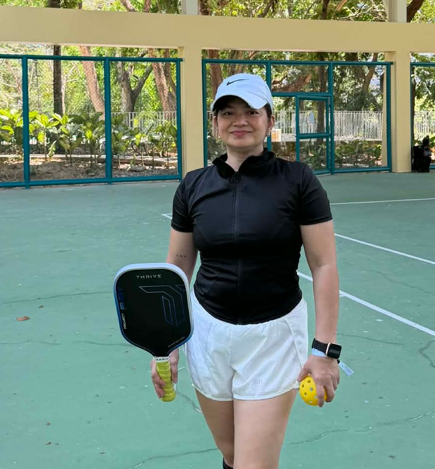 Woman in black shirt and white shorts stands on a green court holding a pickleball paddle and ball, wearing a white cap, with trees in the background.