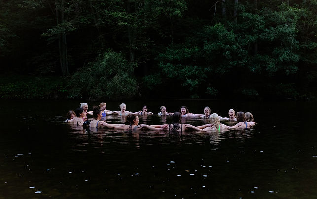 A group of swimmers in a circle, arms linked around each others shoulders, gathered together in the atmospheric waters of a river, surrounded by trees