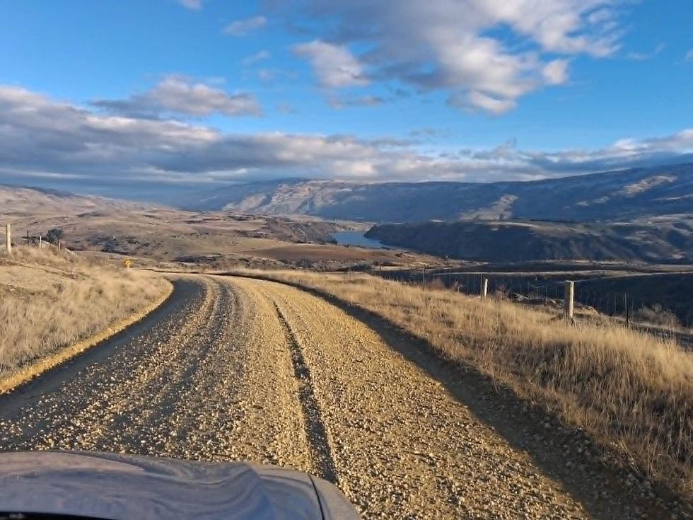 Landscape photo of a gravel road curving through dry hills toward a distant lake and mountains