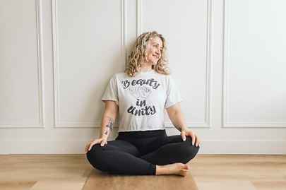 Woman with curly hair sits cross-legged on a mat, smiling in a white room. Shirt reads "Beauty in Unity." Calm and relaxed mood.