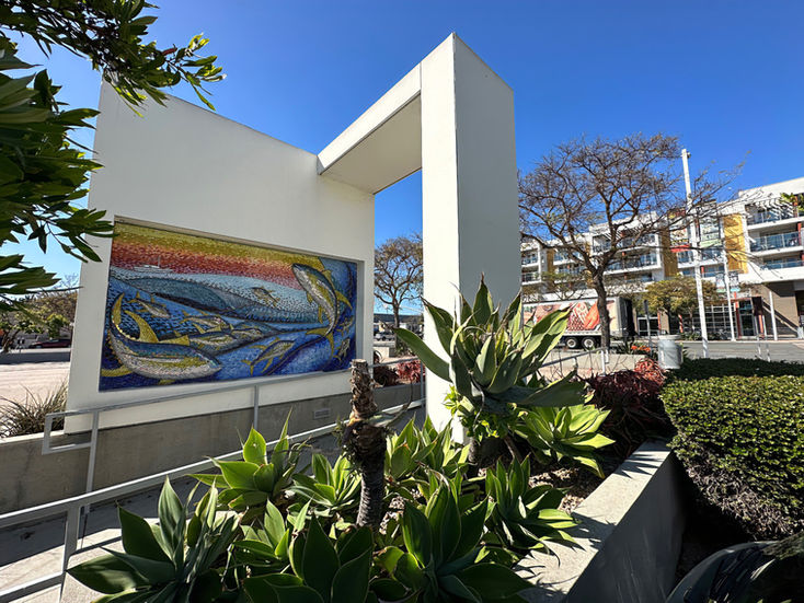 Fish mosaic embedded in white concrete wall with plaza and greenery.