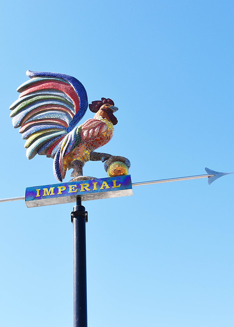 A vibrant mosaic sculpture of a multicolored rooster perched atop a directional weathervane labeled “IMPERIAL” 