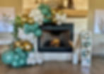 Balloon garland / arch. Colors: Green, white, gold. Baby balloon boxes. Situated around a fireplace