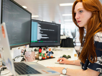 Red-haired woman focuses on coding at dual monitors in a bright office, with code on screens and a laptop on the desk.