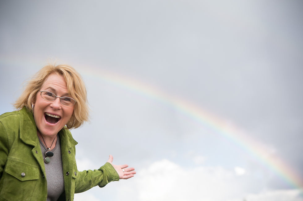 Smiling woman in green jacket gestures under a faint rainbow in a cloudy sky, looking delighted