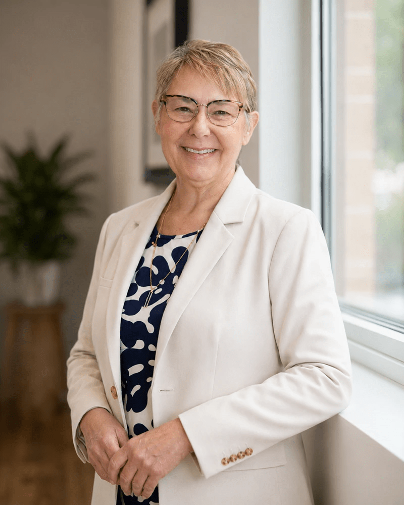 Smiling older woman in glasses and a white blazer stands by a bright window in an office, wearing a navy patterned blouse.