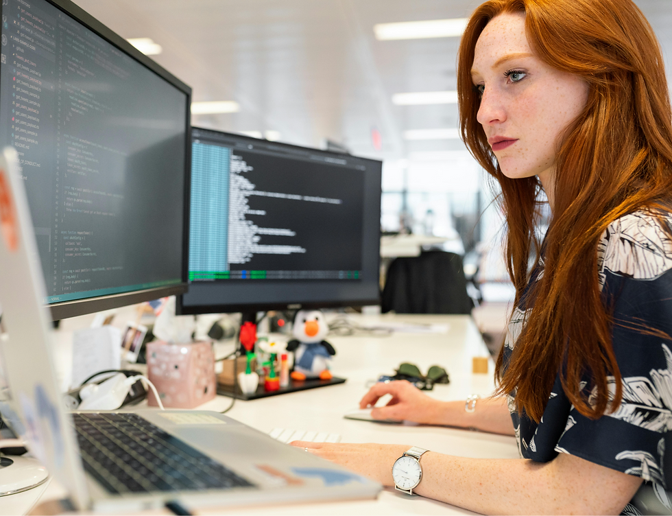 Red-haired woman focuses on coding at dual monitors in a bright office, with code on screens and a laptop on the desk.