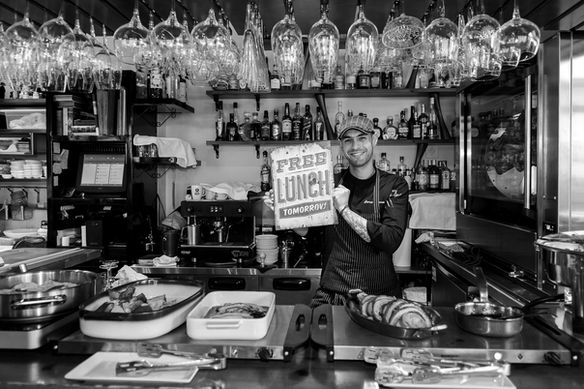 Bartender in a professional bar setting holding a sign that says free lunch tomorrow, black and white image with glasses hanging above and bottles behind, Antonis Karakonstantakis photographer, hospitality industry, restaurant bar, brand personality, candid moment, lifestyle storytelling, service culture