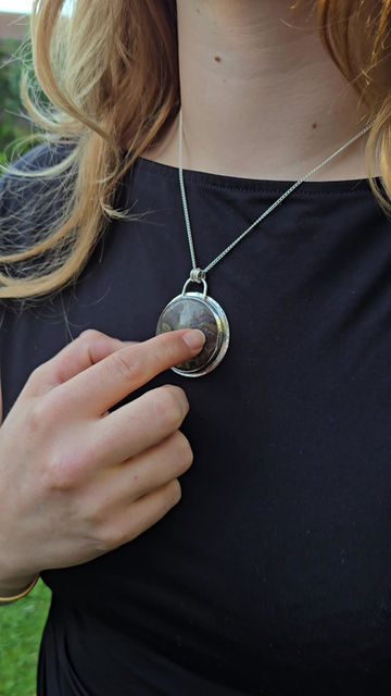 Thumbnail: person wearing a black t-shirt point to silver necklace with circular pendent featuring a dark mottled stone