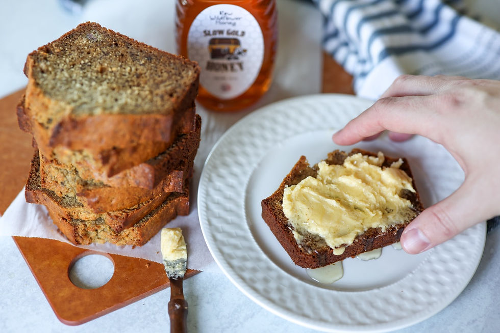 A hand is reaChing into the frame to grab a piece of the best honey banana bread that is topped with bourbon honey butter and drizzled with raw SlowGold Honey. Next to it is a stack of sliced honey banana bread.