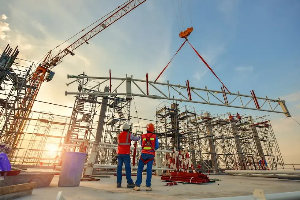 Construction workers in hard hats guide a crane lifting a steel truss at a scaffolded site at sunset.
