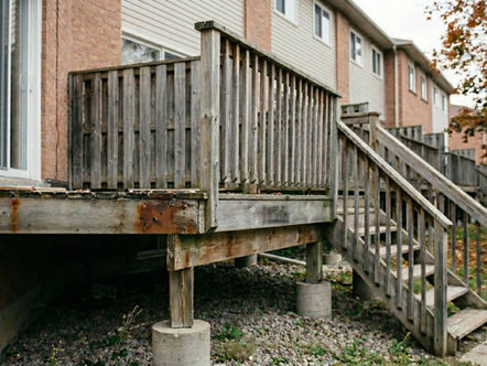 Weathered wooden deck and stairs outside a brick building, surrounded by fallen leaves. Mood is rustic and slightly neglected.