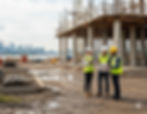 Three construction workers in safety gear discuss blueprints at a muddy site with a city skyline in the background.