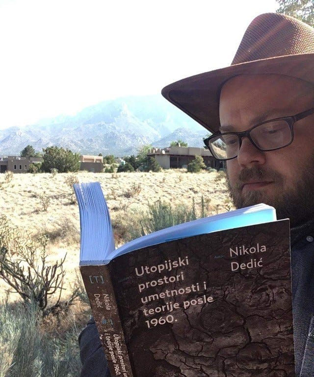 A light-skinned person with a brown hat, glasses, and a beard, reading a book in New Mexico.