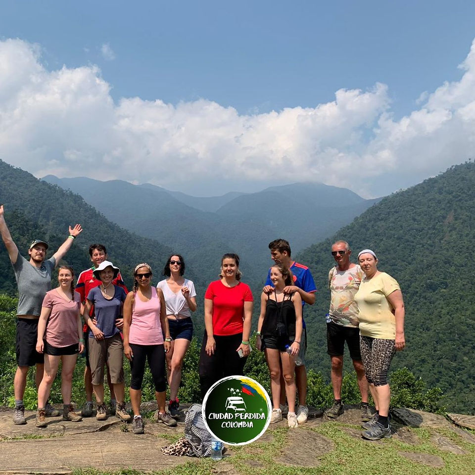 A group of ten hikers poses happily at a viewpoint with a panoramic view of the vast, green mountains of the Sierra Nevada de Santa Marta during the Lost City trek.