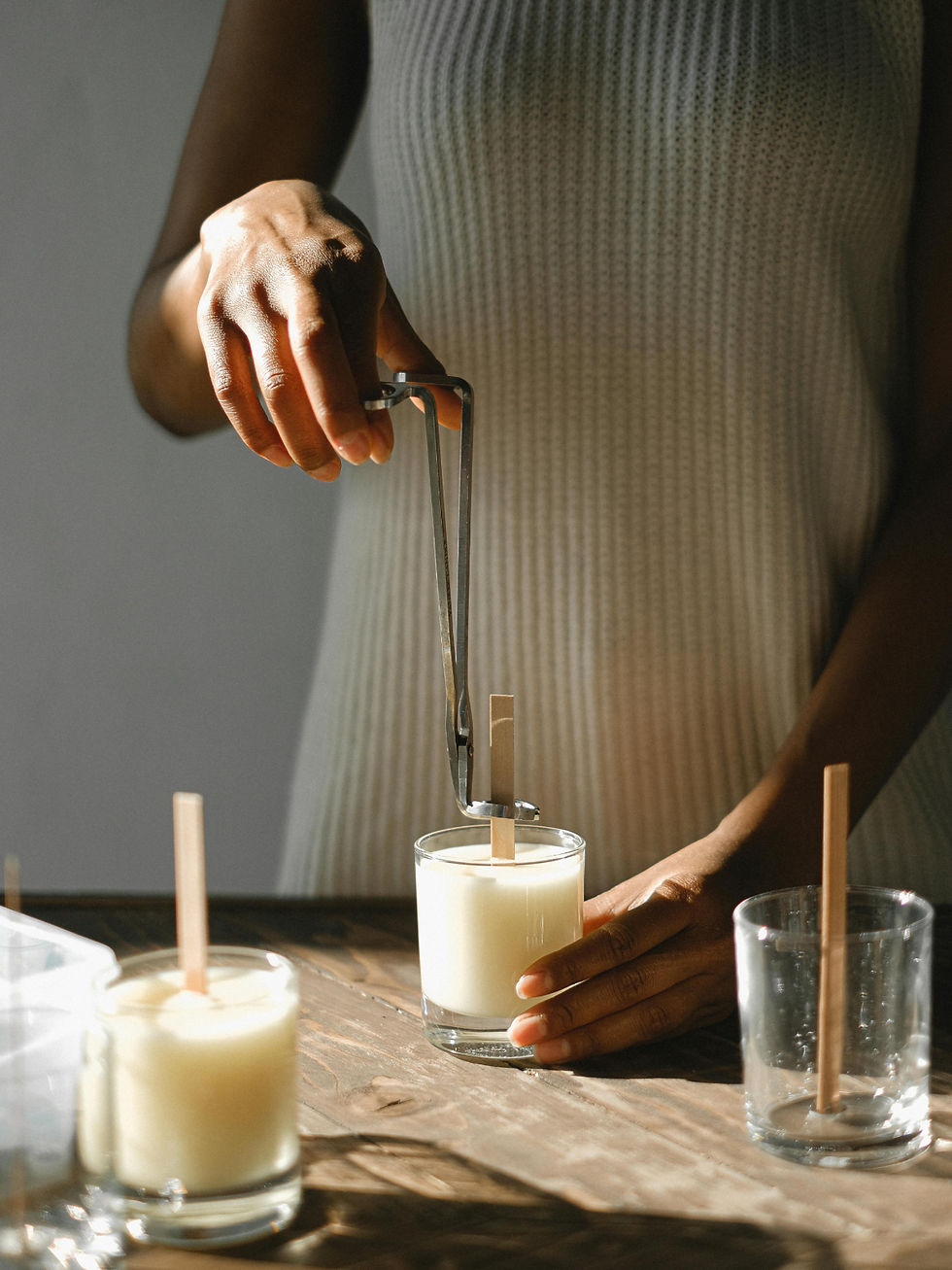 Person in a ribbed dress sets a wick into a white candle glass jar on a sunlit wooden table, with more candles nearby.