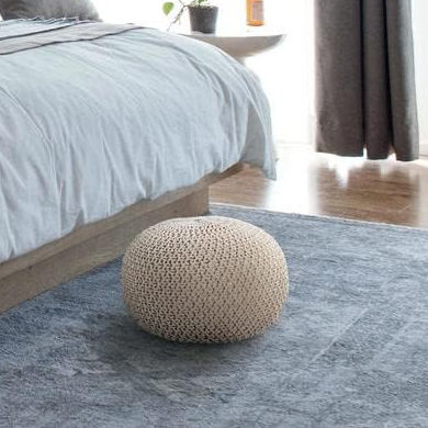 Beige knitted ottoman on blue rug beside a bed with light bedding. Wooden floor, curtains, and small plant in background. Cozy setting.