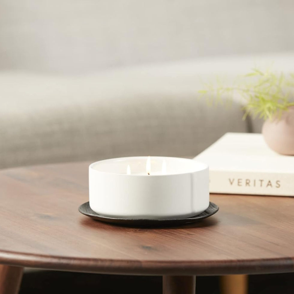A lit white candle on a wooden table beside a book titled "VERITAS" and a potted plant. Cozy and peaceful setting with a beige sofa.