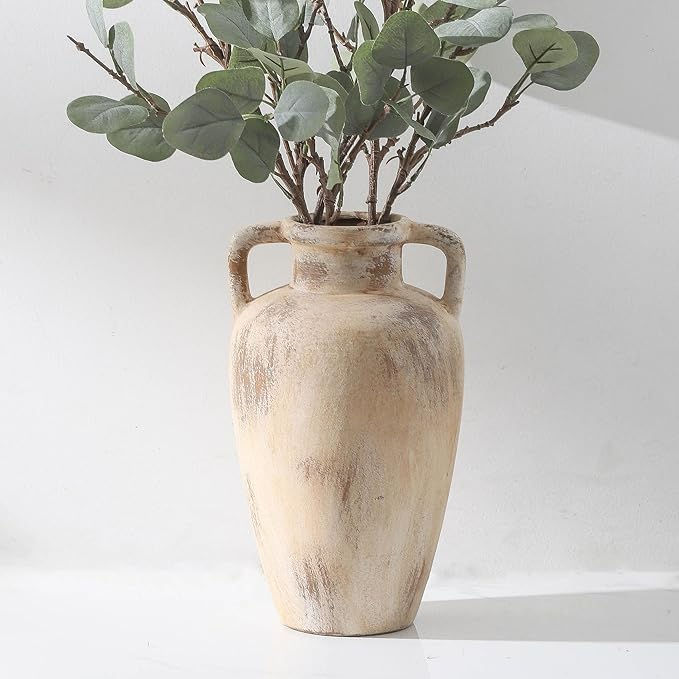 A rustic beige vase with handles holds eucalyptus branches, set against a plain white background. The mood is calm and natural.