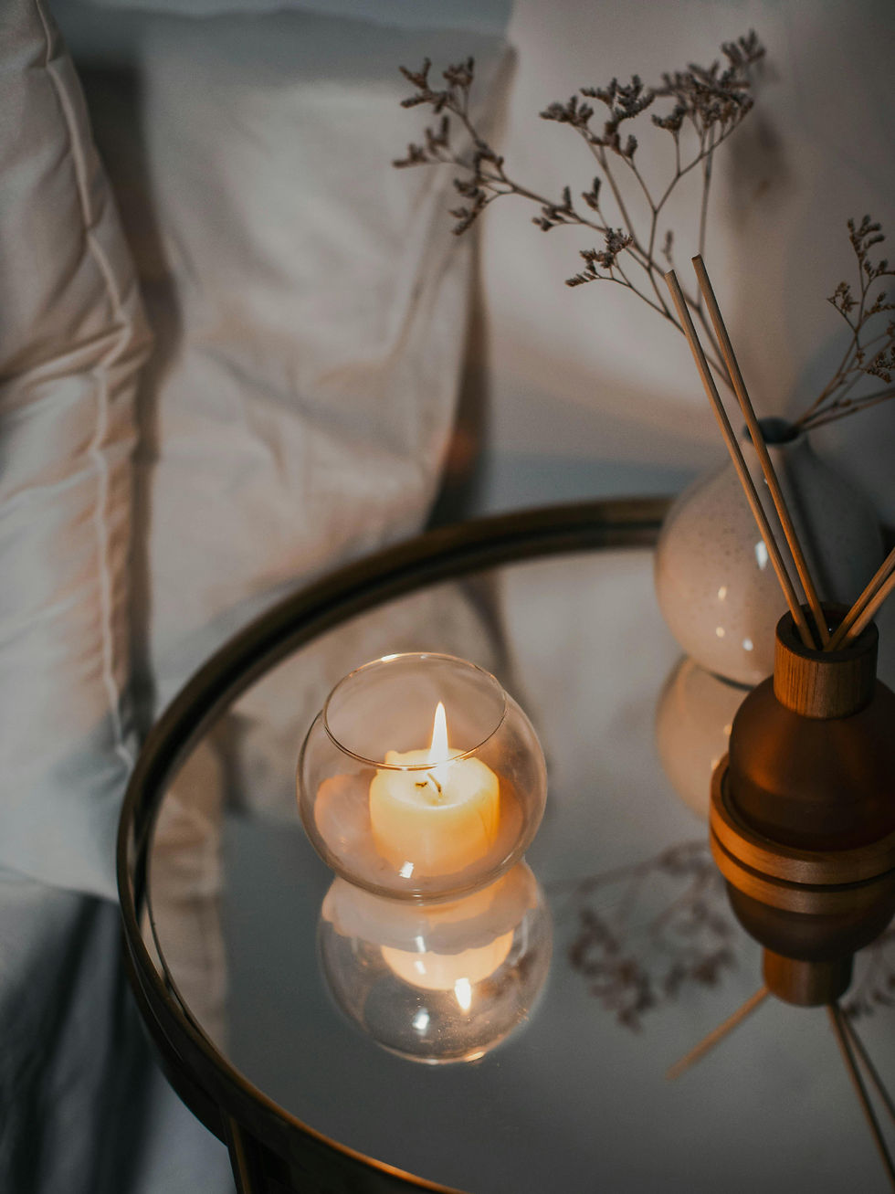 A lit candle in a glass holder sits on a reflective table near a vase with dried branches. Soft, cozy ambiance with neutral tones.