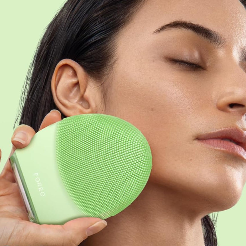 Woman with closed eyes uses green Foreo facial cleansing device on cheek against a pastel green background. Relaxed mood.