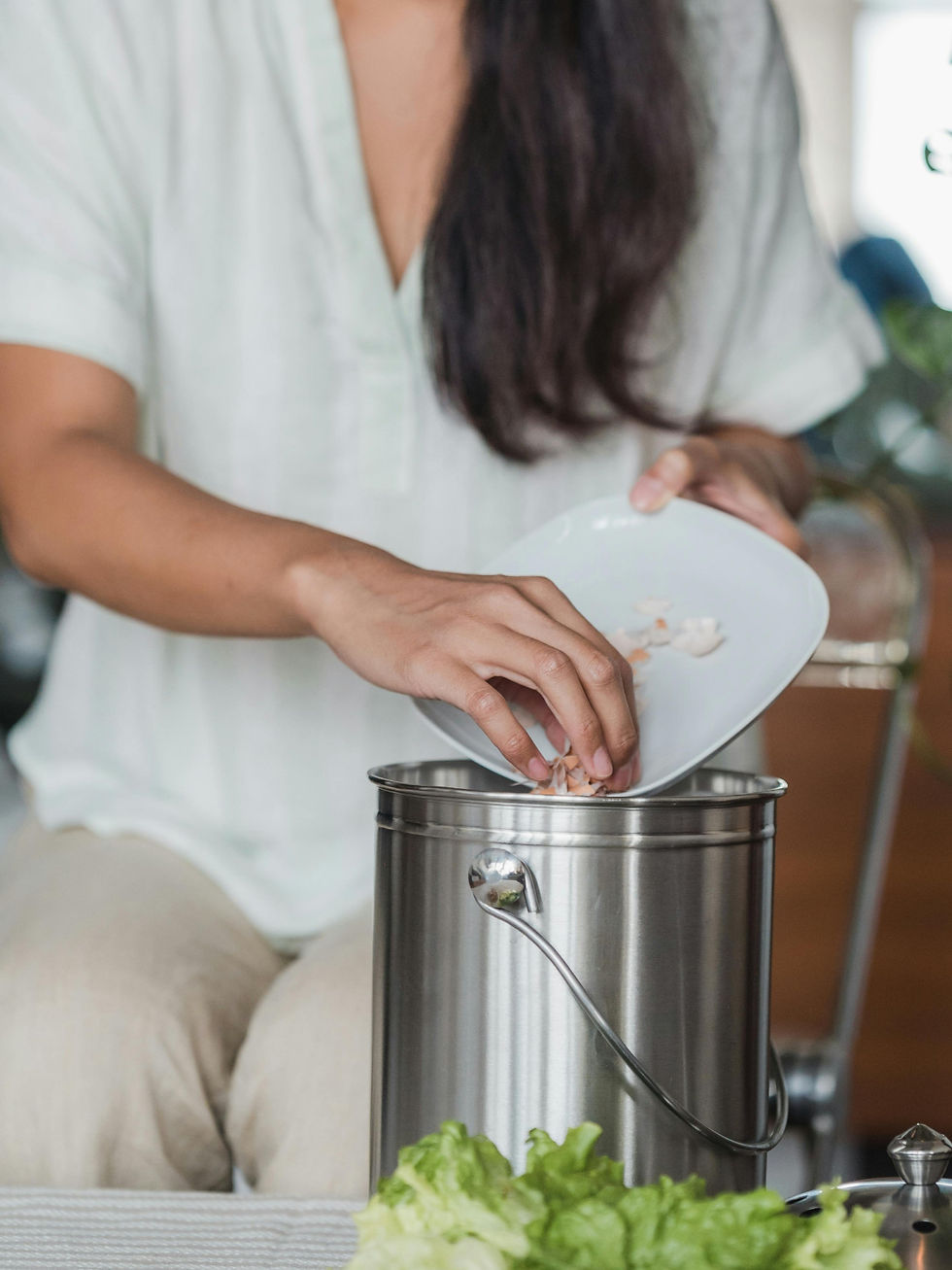 A person in a white shirt empties a plate into a metal compost bin. Green lettuce is visible. Indoor setting with soft lighting.