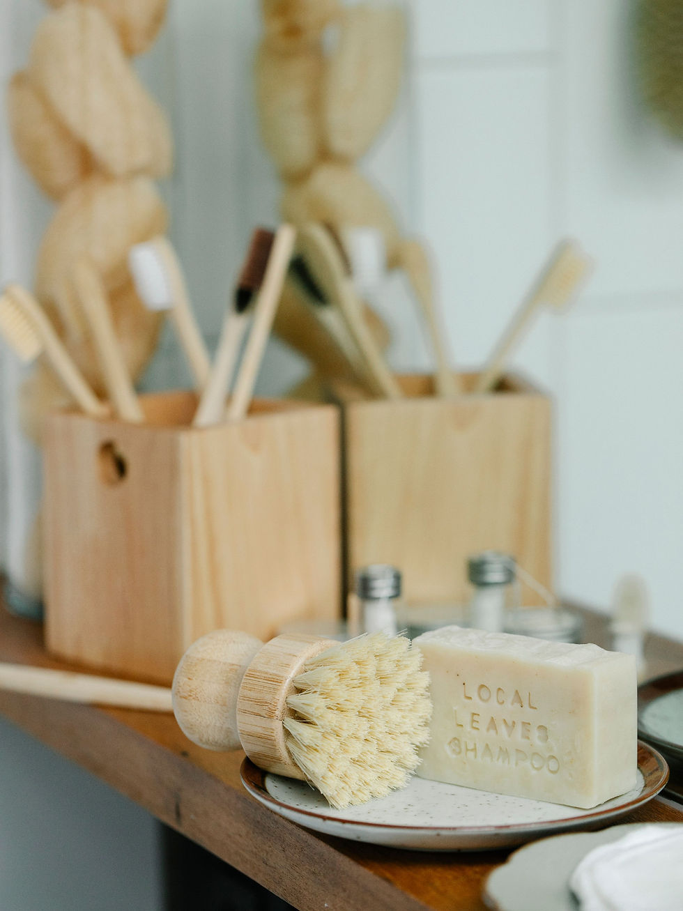Wooden bath brush beside a soap bar embossed LOCAL LEAVES SHAMPOO on a plate, with other wooden brushes in the background.