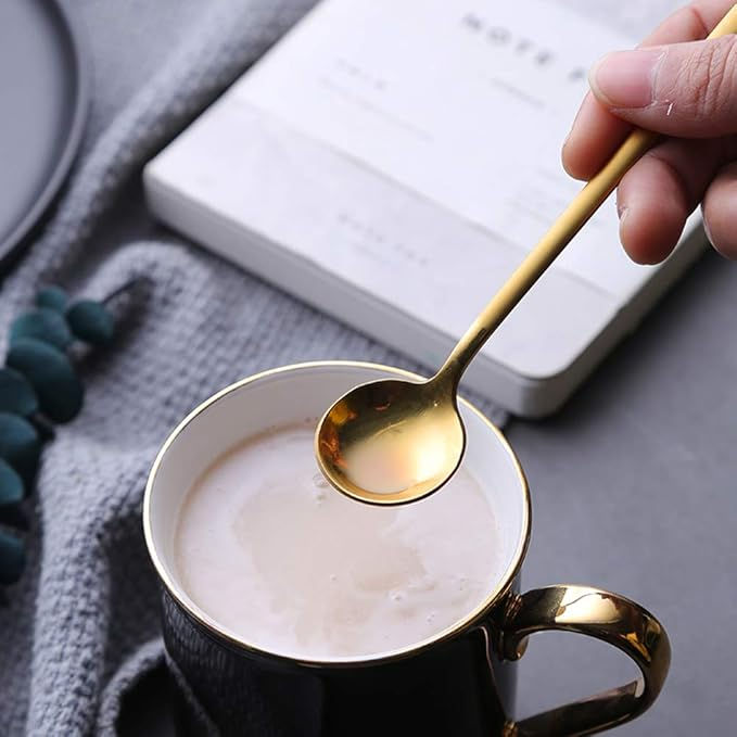 Hand holding gold spoon over hot drink in a black cup. Gray cloth and book on table. Calm, cozy setting.